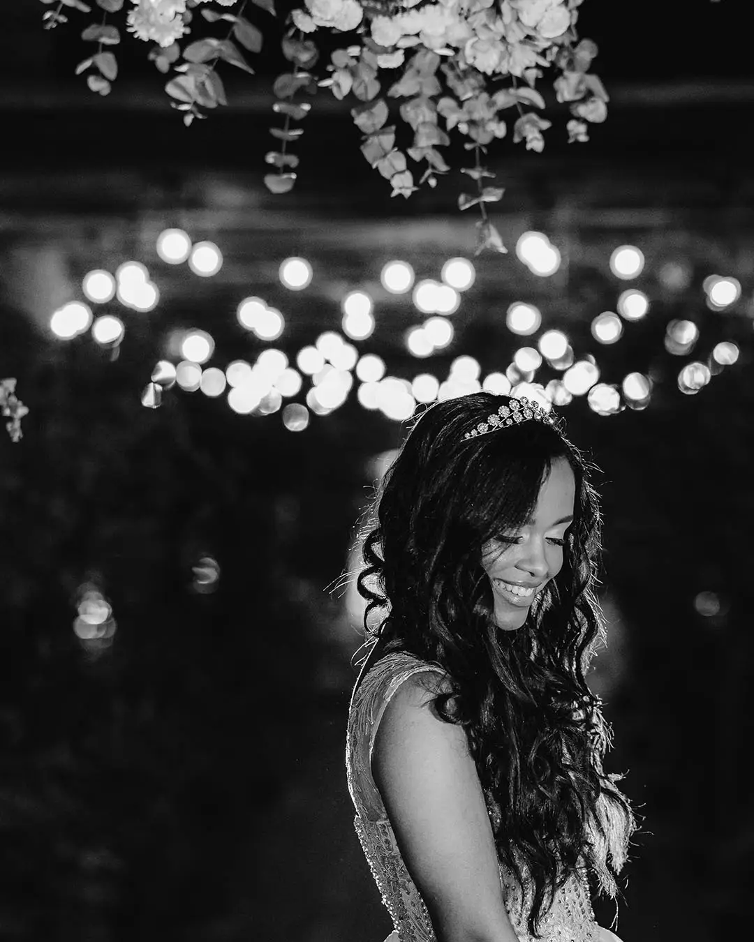 Retrato em preto e branco de uma debutante sorrindo, com tiara, sob luzes desfocadas