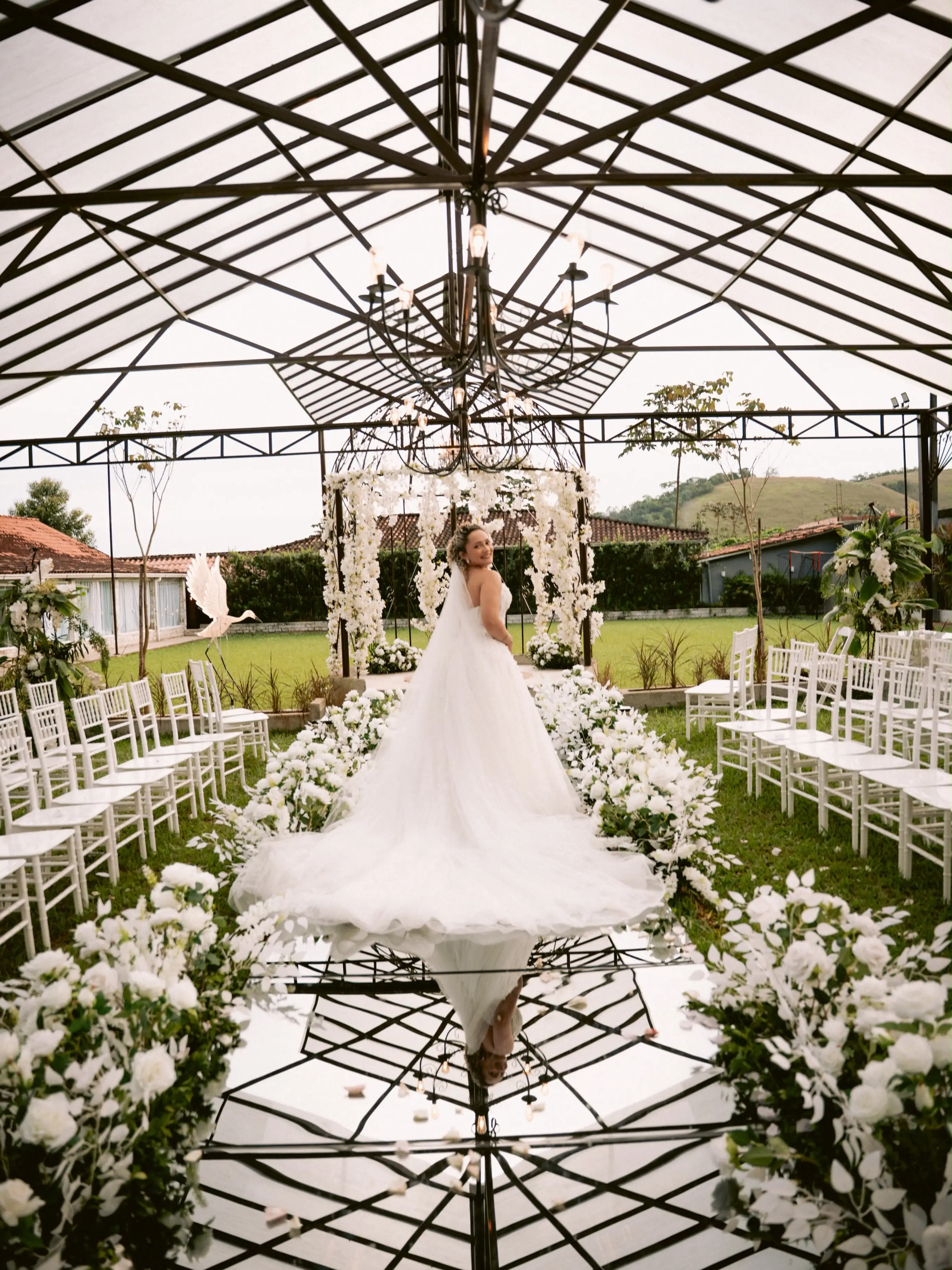 Noiva de costas no corredor da cerimônia, sob cobertura de vidro, entre cadeiras brancas e flores brancas, com a serra ao fundo
