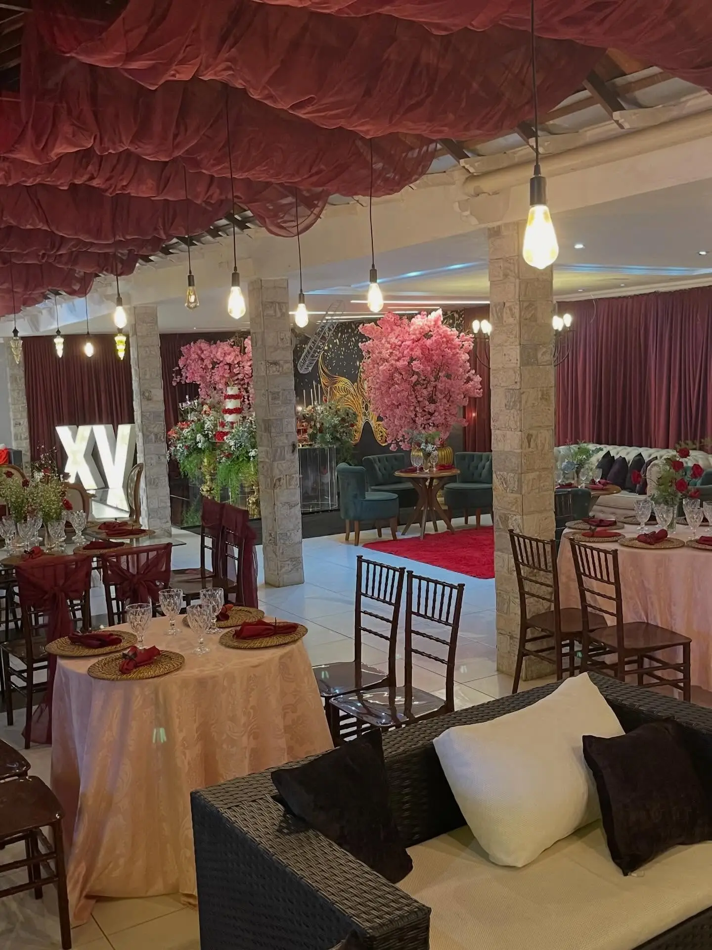 Salão decorado para festa de 15 anos, com letras XV iluminadas, mesas com toalhas rosadas, lounge de sofás e teto drapeado em vinho