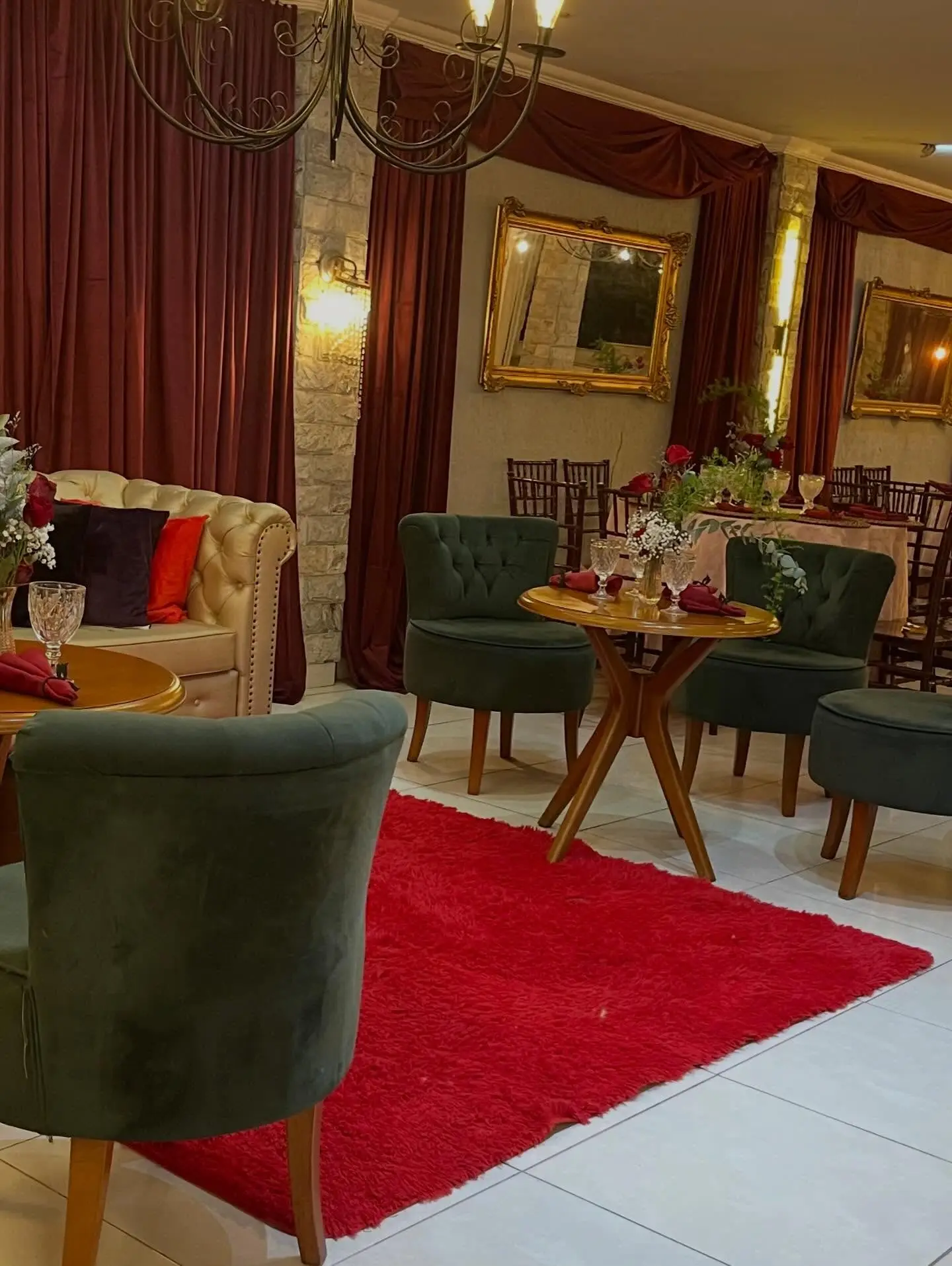 Lounge com poltronas de veludo verde, sofá capitonê dourado, espelho de moldura dourada e tapete vermelho