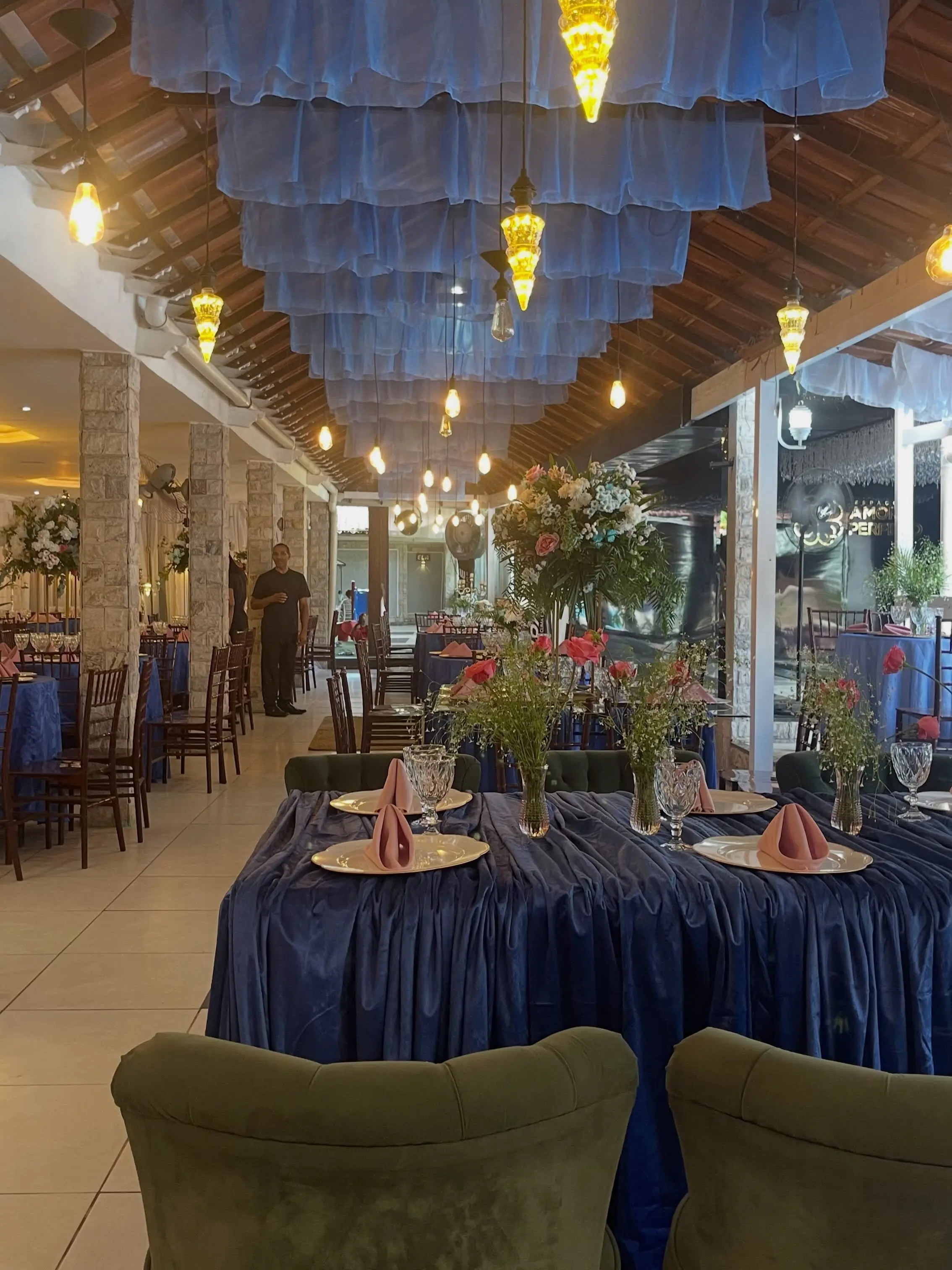 Salão com teto drapeado em tecido azul, luminárias pendentes e mesas postas com toalhas azul-marinho