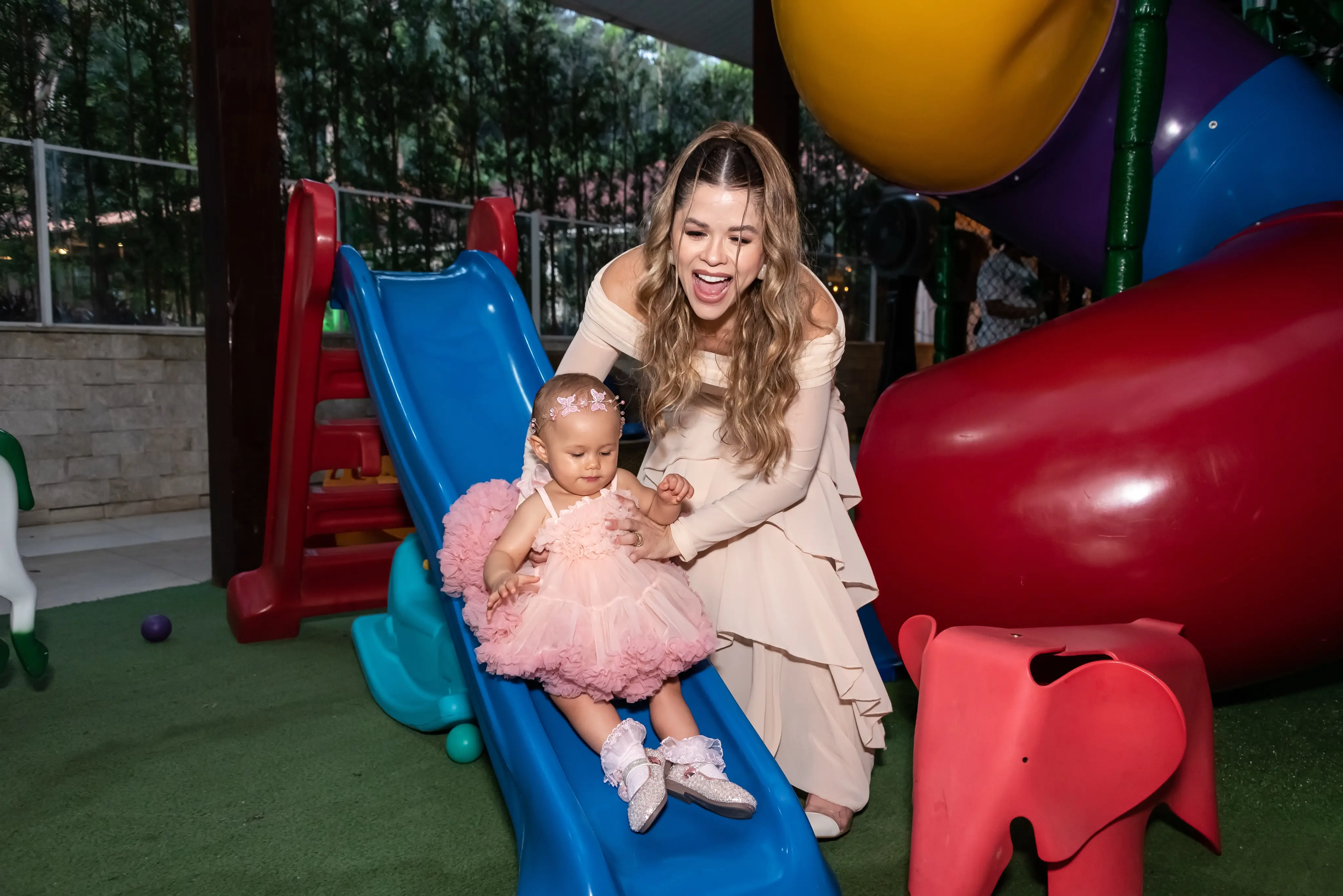 Mãe ajuda a filha pequena, de vestido rosa, a descer um escorregador colorido no gramado