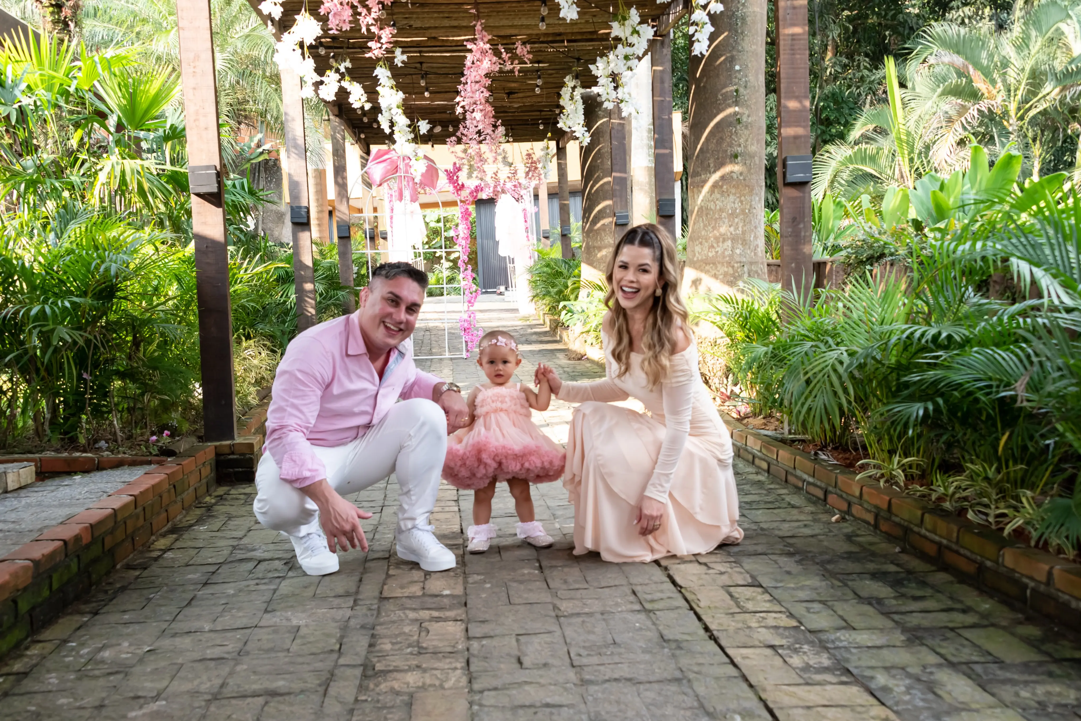 Casal agachado ao lado da filha pequena, de vestido rosa, em um caminho de pedra entre plantas tropicais e arranjos cor-de-rosa