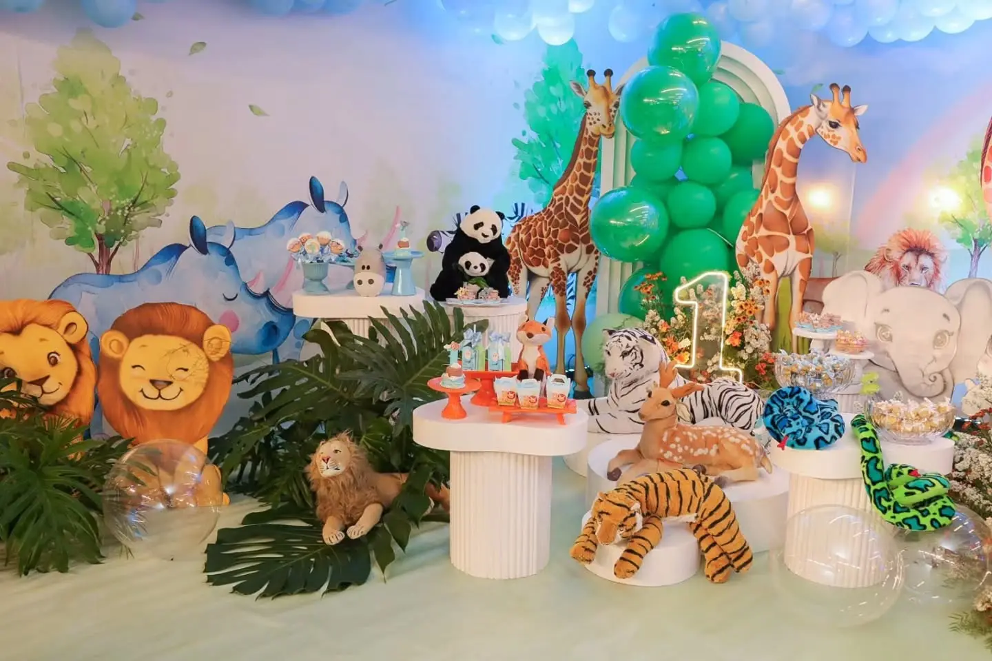 Detalhe da decoração de animais da floresta, com girafas, leões e plantas tropicais ao redor da mesa do bolo