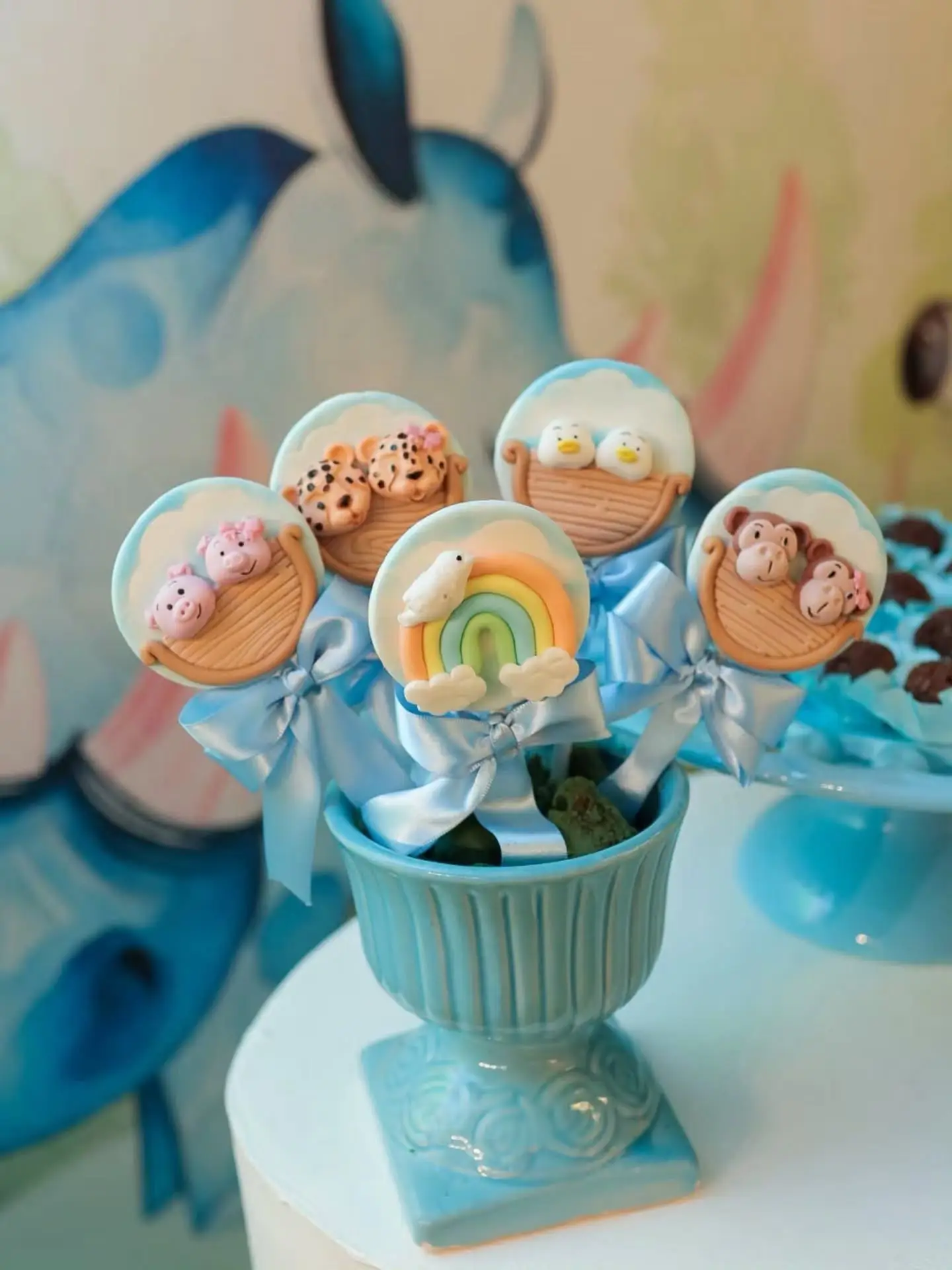 Pirulitos de chocolate decorados com ursinhos e arco-íris, presos com laços azuis em uma taça